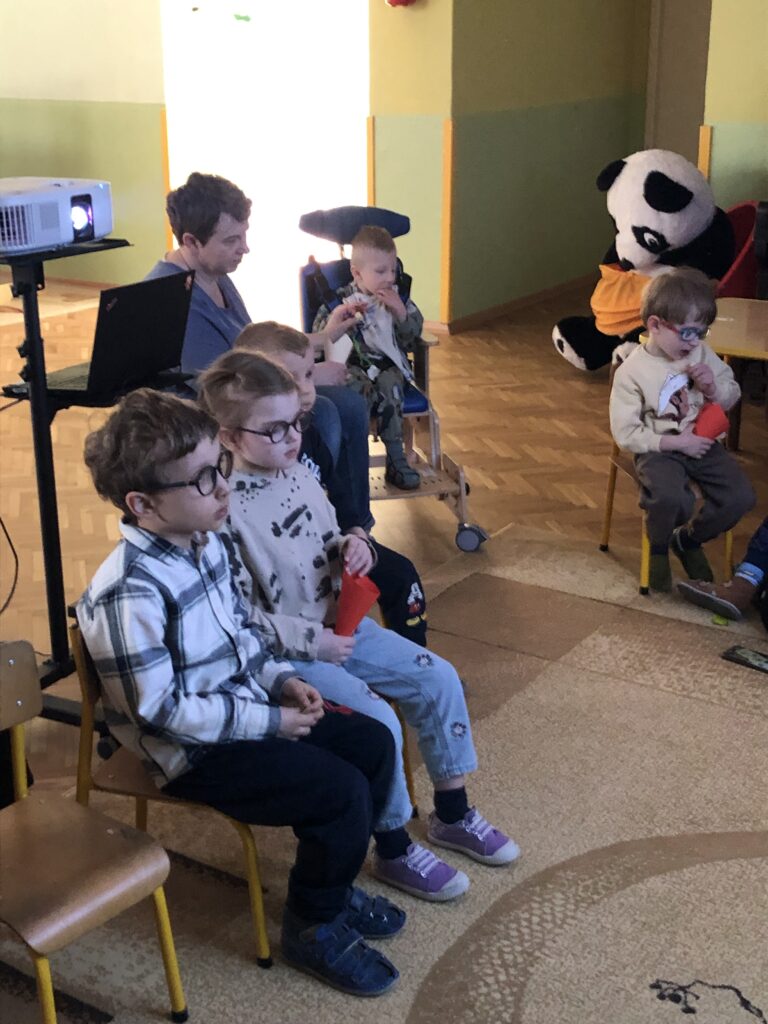 Przedszkolaki siedzą na krzesełkach. Część dzieci trzyma w dłoniach papierowa rożki z chrupkami. Za dziećmi widać stojak z rzutnikiem i laptopem.