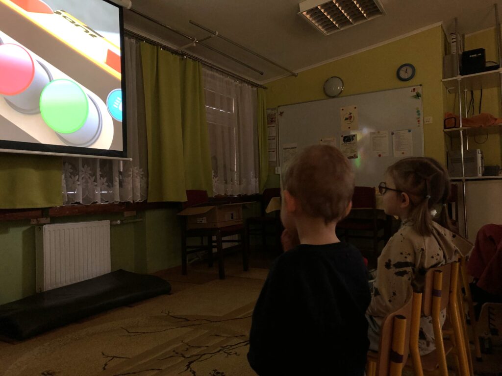 W półmroku dzieci siedzą przed dużym ekranem rzutnika.