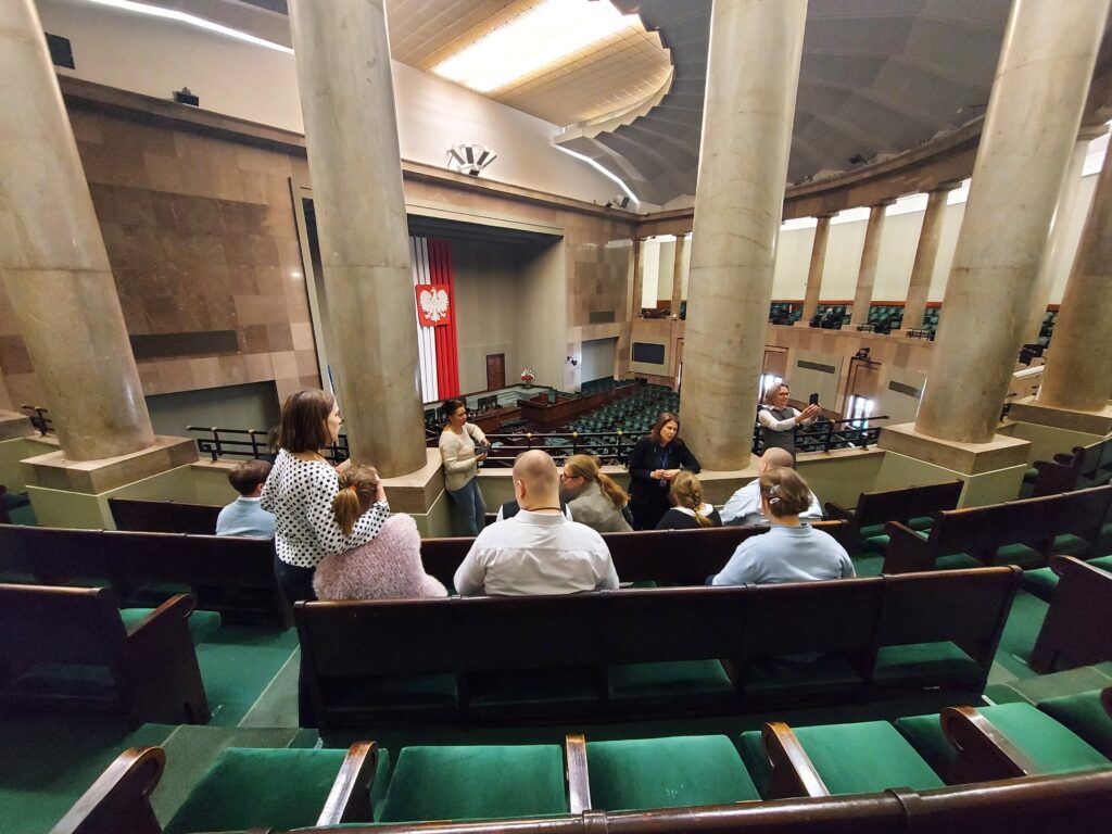 Uczniowie siedzą w galerii Sali Posiedzeń Sejmu i słuchają przewodniczki