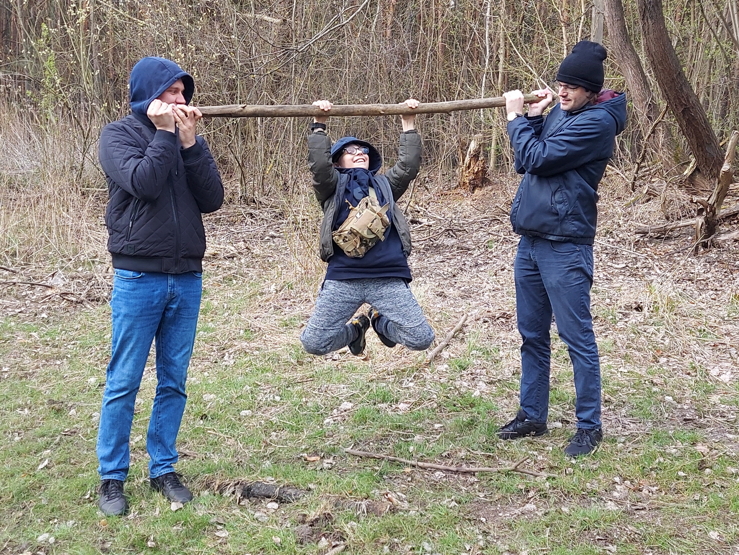dwóch wychowawców trzyma poziomo kij, na którym wisi Piotrek