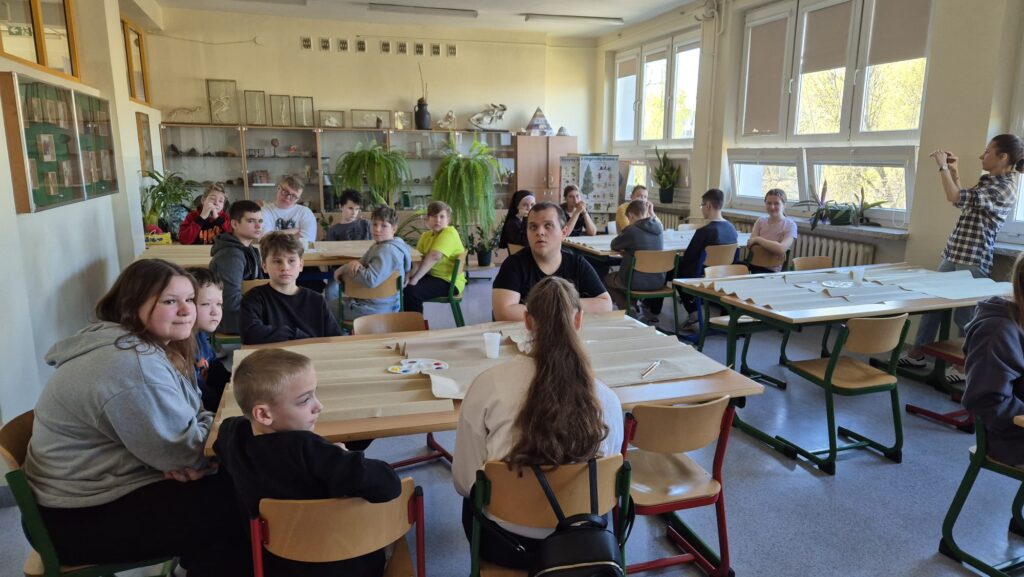 Na zdjęciu widać grupę uczniów siedzących przy stołach w jasnej sali lekcyjnej. Dzieci są skupione,  uczestniczą w zajęciach plastycznych. Na stołach leżą arkusze papieru i farby. W tle widać okna, przez które wpada naturalne światło, oraz szafki z różnymi przedmiotami.