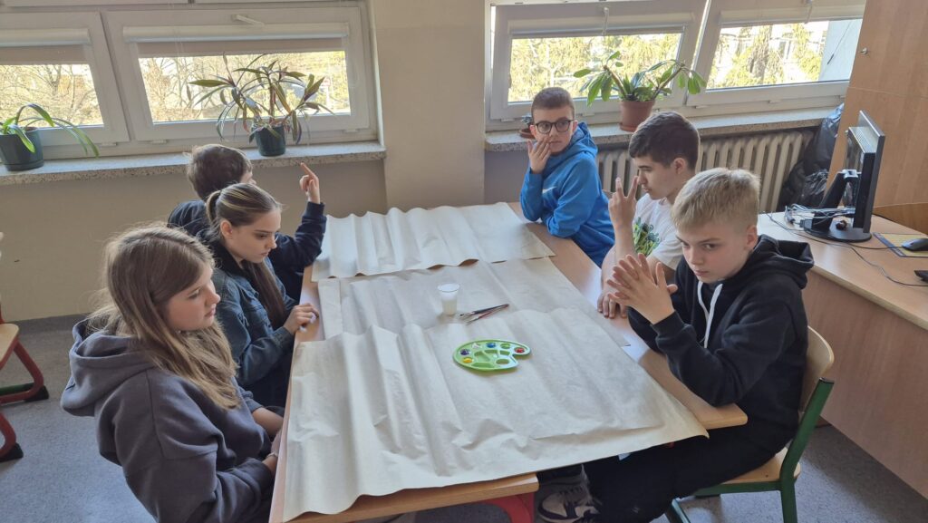 Na zdjęciu widać grupę uczniów siedzących wokół dużego stołu. Na stole rozłożony jest biały papier, a na nim znajduje się paleta z farbami i pędzle, co sugeruje, że dzieci przygotowują się do zajęć plastycznych. 
