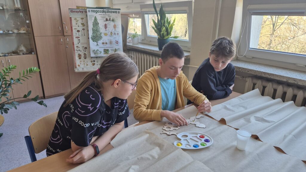 Na zdjęciu widać troje uczniów siedzących przy stole, zajętych pracą nad projektem plastycznym. Na stole znajdują się drewniane elementy, farby i papier, a w tle widoczne są plakaty edukacyjne i okna.