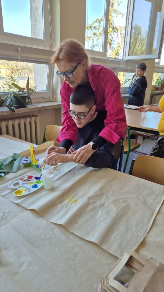 Na zdjęciu widać nauczycielkę, która pomaga uczniowie malować drewnianą ozdobę. 