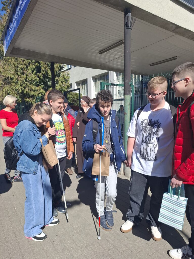 Na zdjęciu widać grupę uczniów stojących na chodniku przed budynkiem. Dwoje uczniów trzyma białe laski. Pozostali uczniowie trzymają papierowe torby.