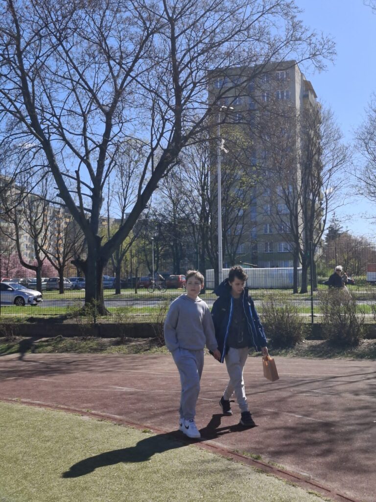 Na zdjęciu widać dwóch uczniów idących chodnikiem w parku. Trzymają się za ręce, a jeden z nich trzyma papierową torbę. W tle widać budynki mieszkalne i drzewa bez liści.
