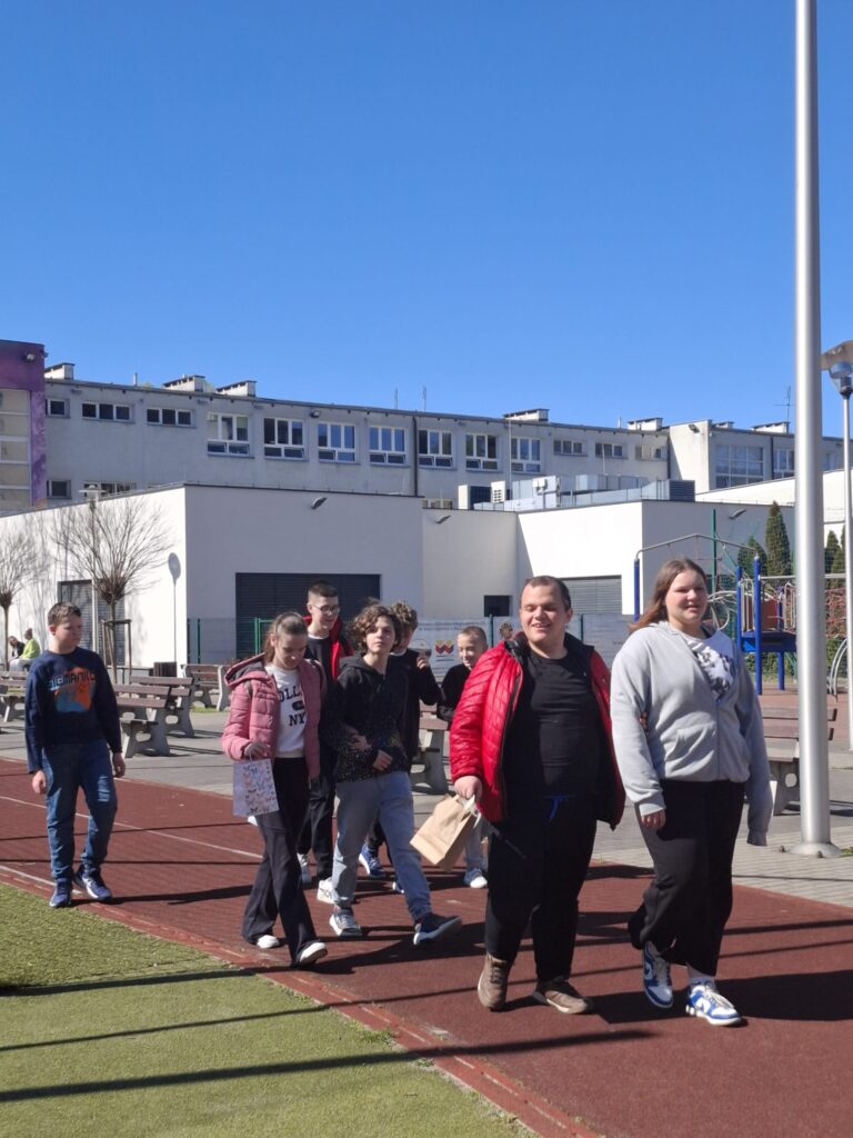 Na zdjęciu widać grupę uczniów idących po bieżni lekkoatletycznej. Uczniowie są uśmiechnięte i trzymają w rękach papierowe torby. W tle widać budynek szkoły i niebieskie niebo.