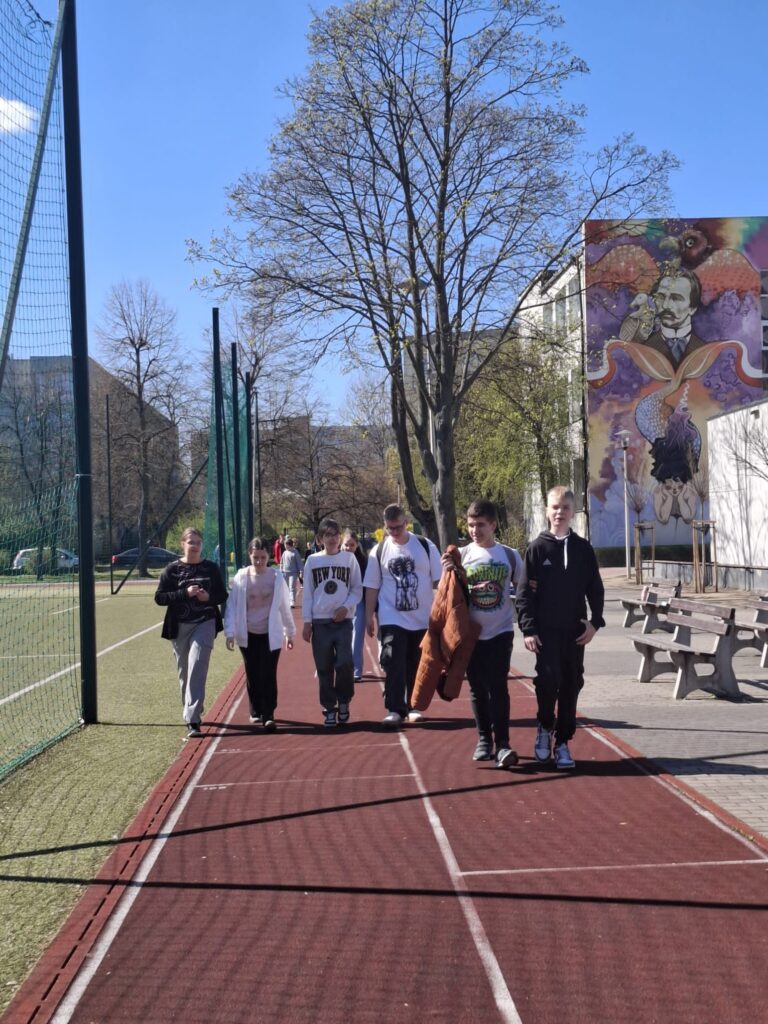 Na zdjęciu widać grupę uczniów idących po czerwonej bieżni lekkoatletycznej. Dzieci zmierzają w kierunku budynku szkoły, na którego ścianie widać duży obraz.