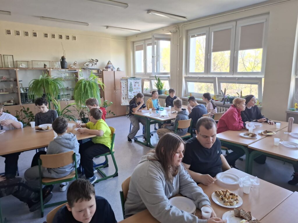 Na zdjęciu widać grupę uczniów siedzących przy stołach w klasie i jedzących posiłek. Uczniowie są skupione na jedzeniu, a na stołach leżą talerze z jedzeniem.