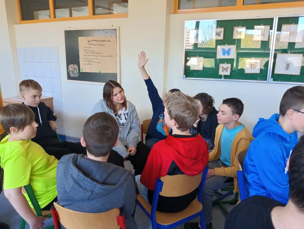 Na zdjęciu widać grupę uczniów siedzących w kręgu w klasie. Jedn z uczniów trzyma podniesioną rękę, jakby chciała zadać pytanie lub zabrać głos. W tle widać tablicę z rysunkami owadów i motyli.