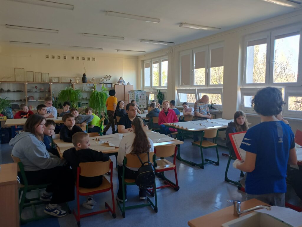 Na zdjęciu widać grupę uczniów siedzących przy stołach w klasie, którzy uczestniczą w lekcji. Na stołach leżą arkusze papieru i przybory szkolne.