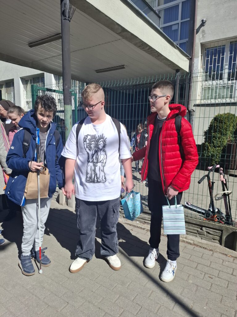 Na zdjęciu widać trzech uczniów stojących na chodniku przed szkołą. Jeden z chłopaków trzyma białą laskę. 
