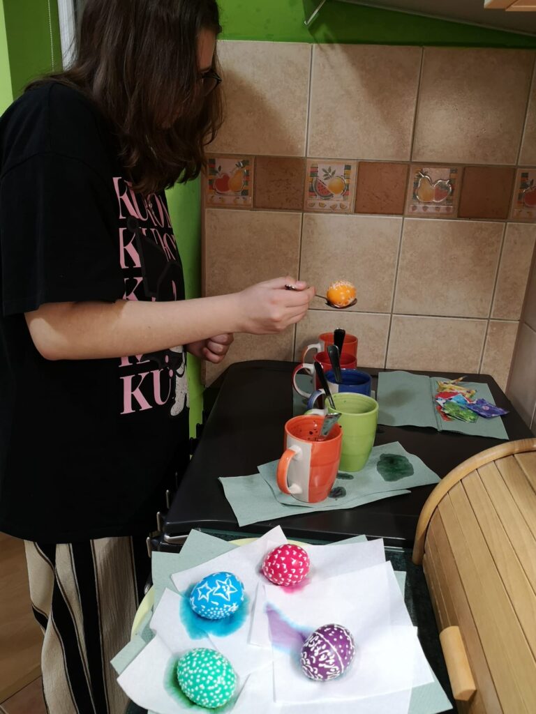Ania wyjmuje z barwnika jajko na dużej łyżce, jajko umalowało się na kolor pomarańczowy obok leżą kraszanki w kolorze niebieskim, zielonym, fioletowym i czerwonym.