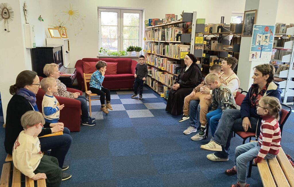 Nauczyciele i dzieci siedzą w kole na krzesłach i ławkach. Za nimi widać pianino, kanapę i regały z książkami. 