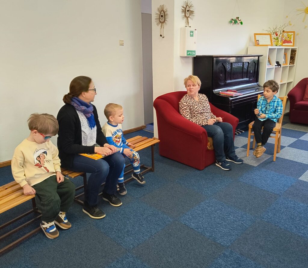 Na zdjęciu nauczycielka z dwoma chłopcami siedzi na ławce. Obok pani bibliotekarka siedzi na fotelu i chłopiec na krześle. Za nimi widać pianino i regał na książki.