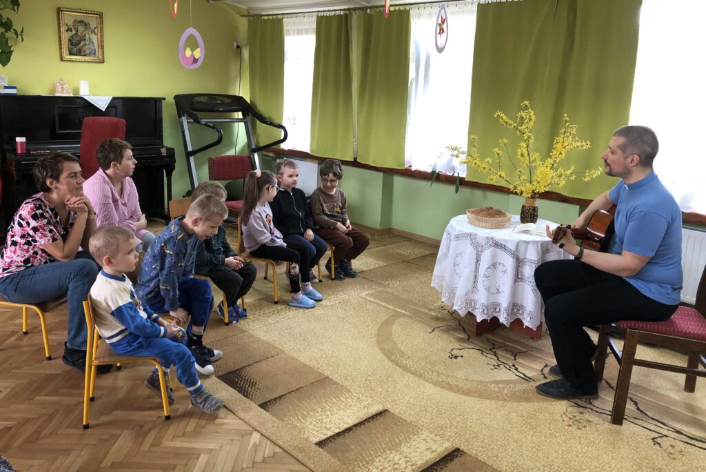 Dzieci i nauczyciele siedzą na krzesełkach. Na przeciwko siedzi ksiądz z gitarą. Pomiędzy nimi stoi stolik nakryty białym obrusem, a na nim chleb, biblia i forsycja w wazonie.