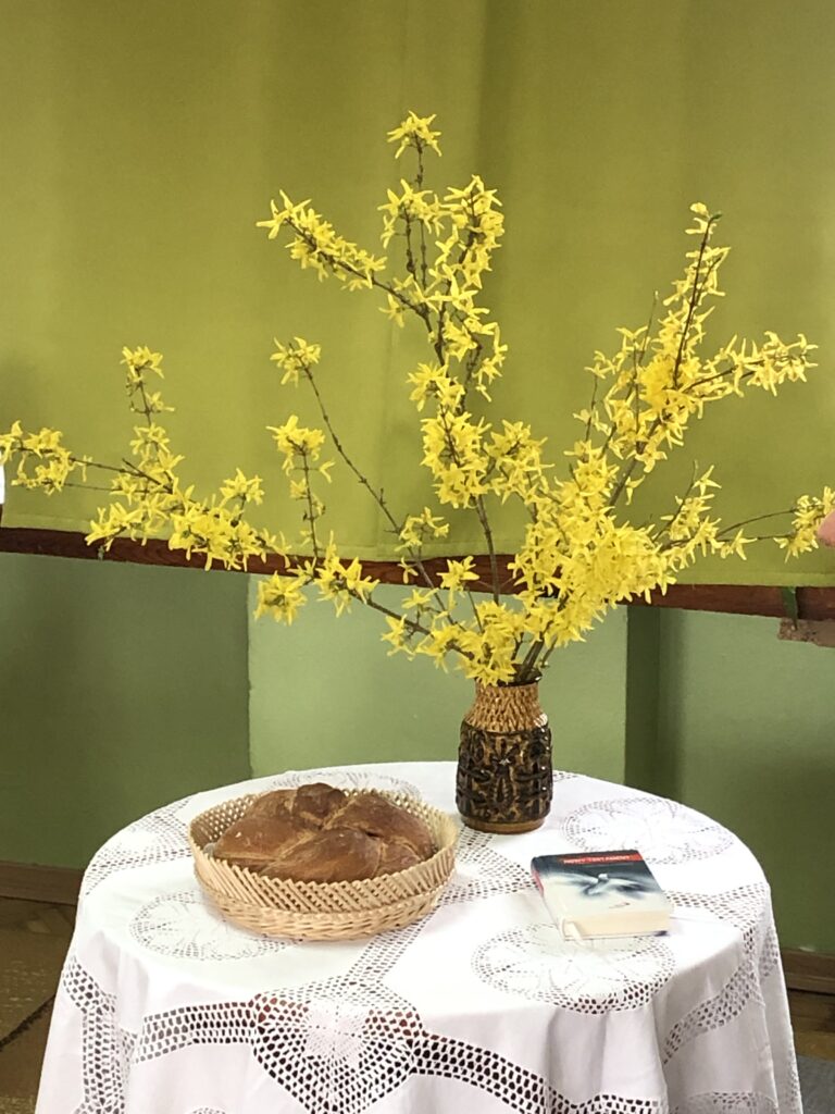 Okrągły stolik nakryty białym obrusem. Na stoliku koszyk z okrągłym bochenkiem chleba, Pismo Święte i forsycja w wazonie.
