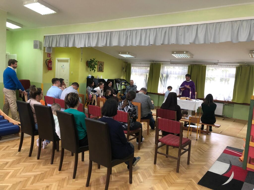 Rodzice, nauczyciele i dzieci siedzą na krzesłach na bawialni przedszkolnej. Ksiądz stoi przy ołtarzu. 