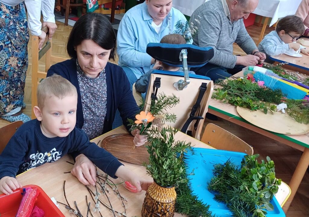 Na pierwszym planie chłopiec przy stoliku sięga po gałązki bukszpanu. Obok widać nauczycieli i rodziców z dziećmi robiących palemki wielkanocne.