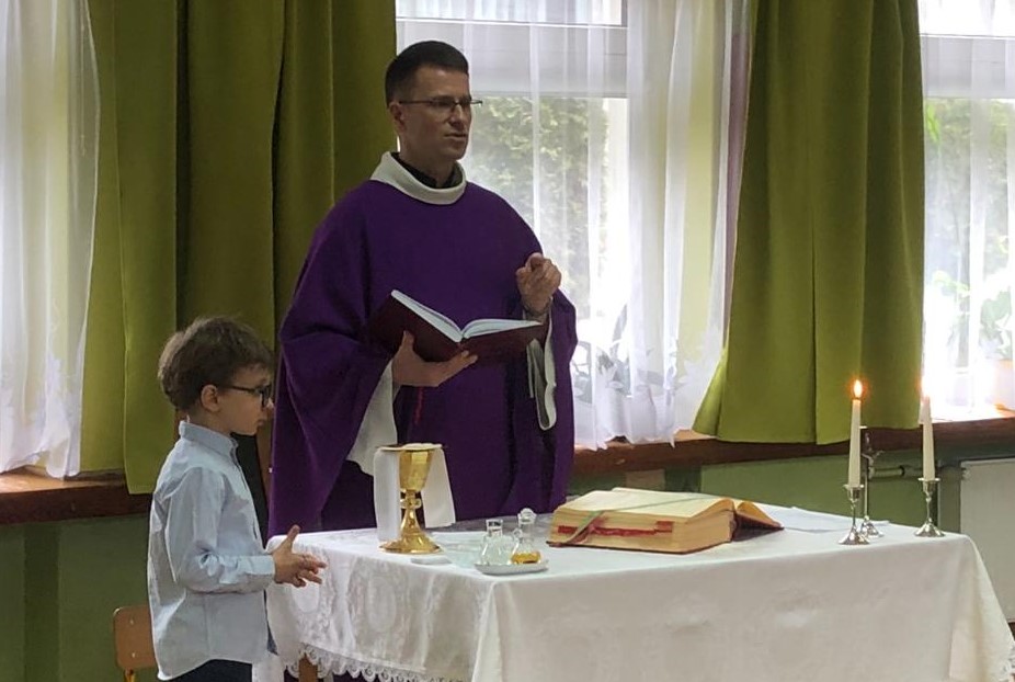 Ksiądz z otwartym lekcjonarzem w dłoni stoi przy ołtarzu na bawialni przedszkolnej. Obok stoi chłopiec.