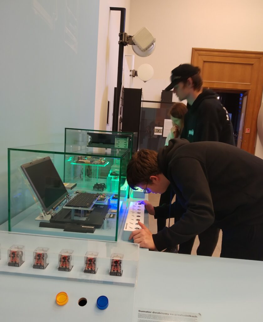 Trzy osoby, dwie z nich to uczniowie, a jedna to kobieta, oglądają interaktywną ekspozycję w muzeum lub galerii. Na pierwszym planie widać szklaną gablotę z otwartym laptopem i innymi elementami elektronicznymi, a także panel sterujący, którym manipuluje jeden z uczniów.
