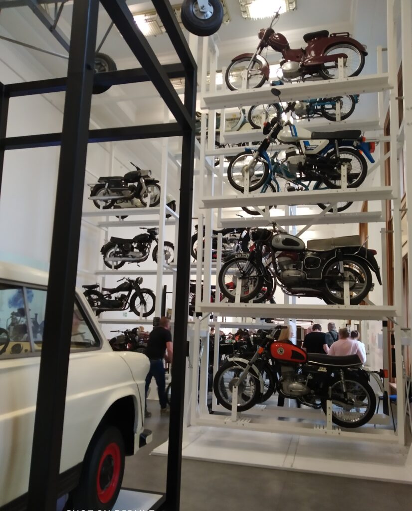 Zdjęcie przedstawia wnętrze muzeum, prawdopodobnie Narodowego Muzeum Techniki w Warszawie, gdzie na metalowych regałach i na parterze wystawionych jest wiele zabytkowych motocykli. Na pierwszym planie widoczny jest fragment białego samochodu, a w tle można dostrzec kilku zwiedzających.