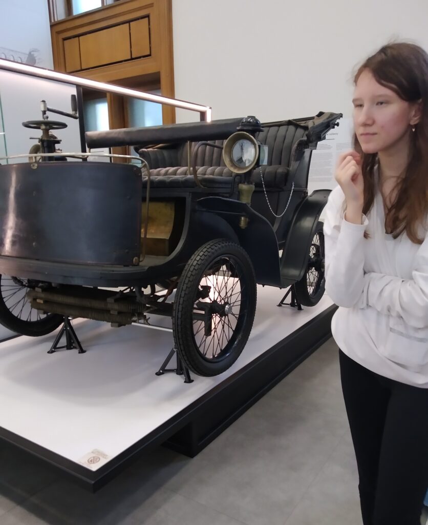 Zapytanie zostało przetworzone
Na zdjęciu widoczny jest zabytkowy samochód, prawdopodobnie Peugeot Type 19, wystawiony w muzeum. Obok pojazdu stoi uczennica z długimi włosami, zamyślona, patrząca w prawo.