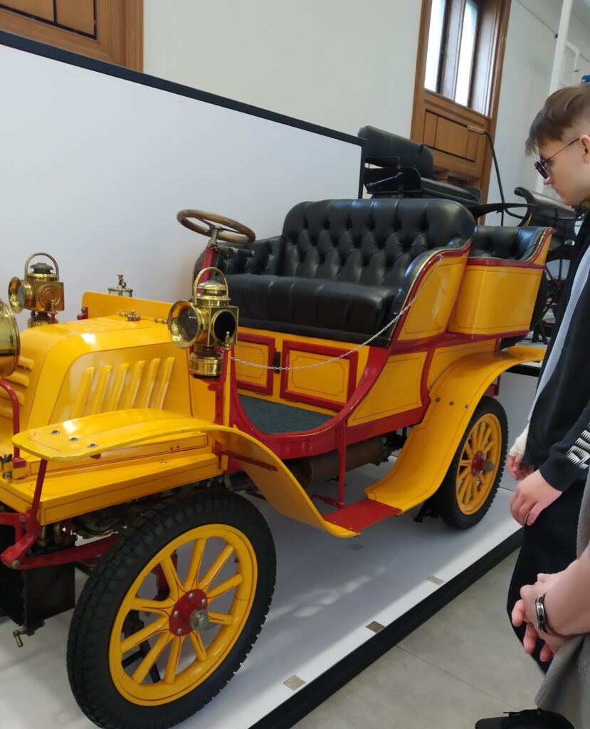 Zdjęcie przedstawia zabytkowy żółty samochód, prawdopodobnie model Benz Spider z 1900 roku lub Adler z 1901 roku, z czarną skórzaną tapicerką. Po prawej stronie kadru widoczna jest część osoby w okularach, która ogląda eksponat.