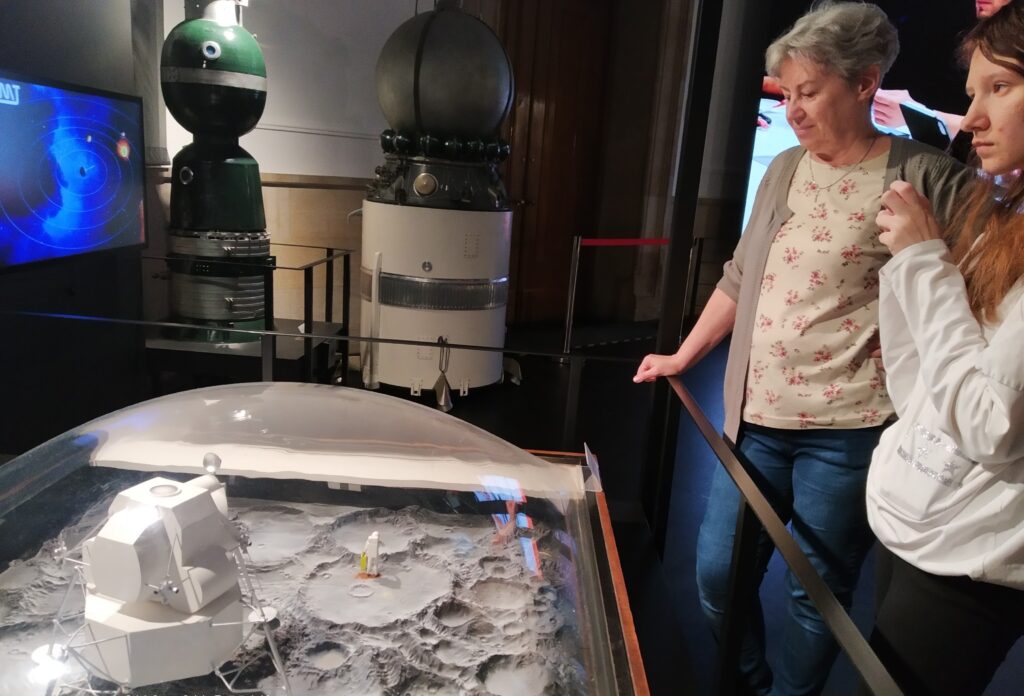 Zapytanie zostało przetworzone
Zdjęcie przedstawia dwie osoby, kobietę i młodą osobę, oglądające wystawę w muzeum kosmicznym. Na pierwszym planie widać makietę powierzchni księżyca z lądownikiem i figurką astronauty, a w tle repliki statków kosmicznych i ekrany wyświetlające informacje.