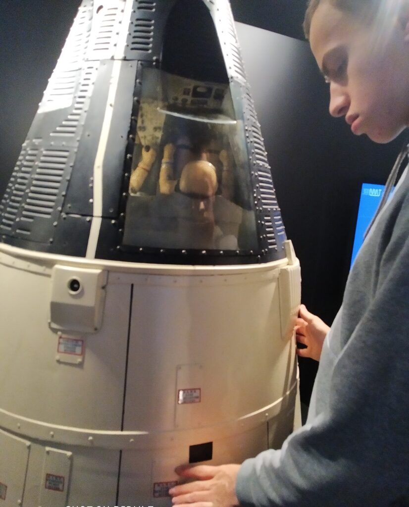 Uczeń technikum dotyka dłońmi eksponatu, który wygląda jak kapsuła kosmiczna, znajdującego się w muzeum. Przez okno kapsuły widać odbicie innej osoby.