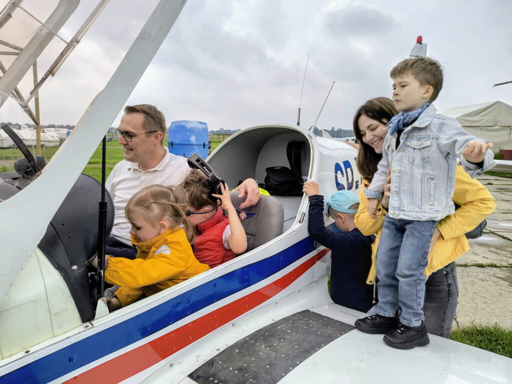 Dzieci siedzące z pilotem w kabinie samolotu. Chłopiec stojący na skrzydle , za nim kobieta