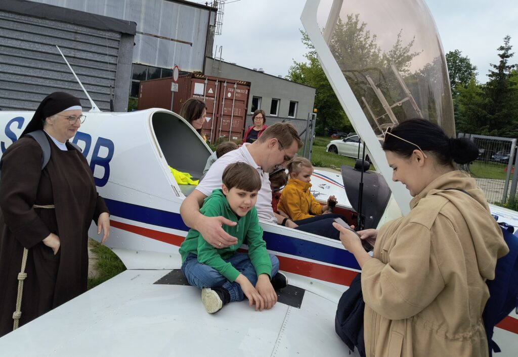 Chłopiec siedzący na skrzydle samolotu. Za nim pilot z dziewczynką w kabinie. Dwie osoby stojące po bokach skrzydła. 
