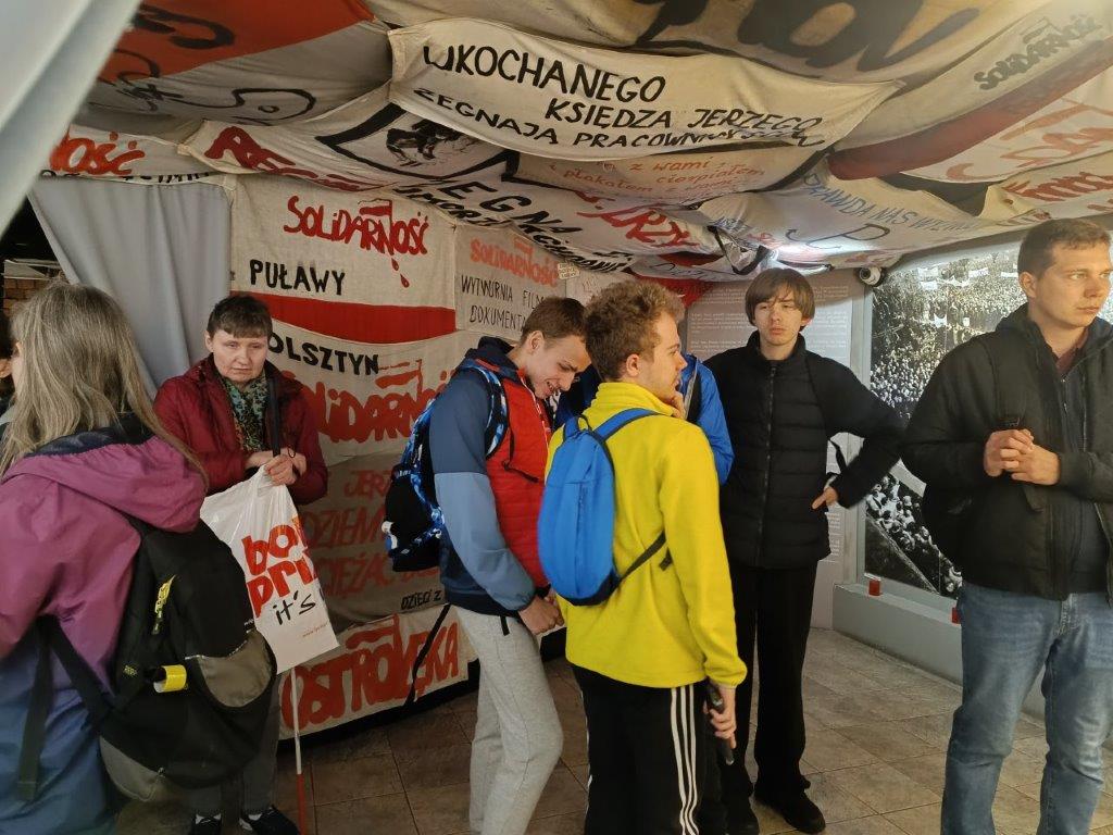 Grupa zwiedza Muzeum Błogosławionego Księdza Jerzego Popiełuszki, znajdując się w pomieszczeniu z transparentami "Solidarności" i gablotami z eksponatami, co wskazuje na wystawę poświęconą historii tego okresu.