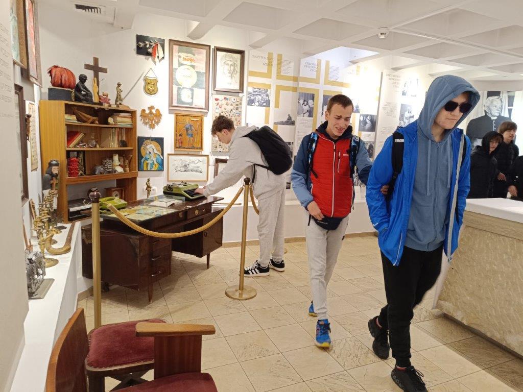 Trzech uczniów zwiedza Muzeum Błogosławionego Księdza Jerzego Popiełuszki, oglądając liczne eksponaty, w tym obrazy, pamiątki osobiste i zabytkowe meble z maszyną do pisania.