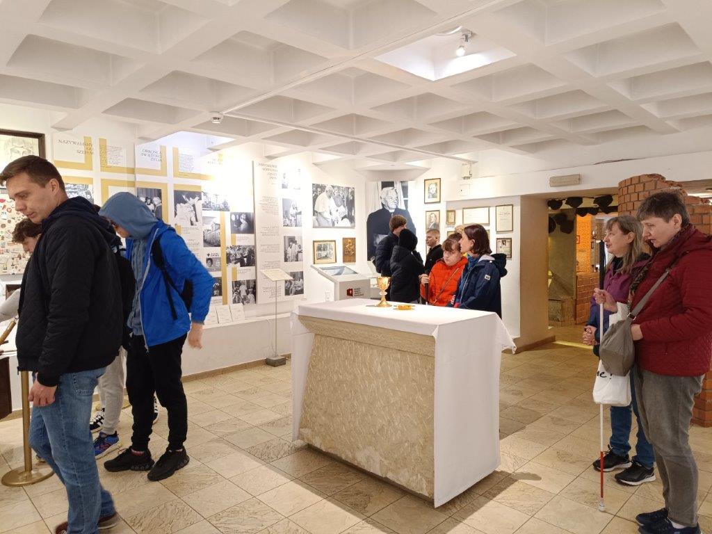 Na zdjęciu widać wnętrze Muzeum Błogosławionego Księdza Jerzego Popiełuszki, gdzie grupa zwiedzających ogląda wystawione eksponaty. W centralnej części pomieszczenia, pod kasetonowym sufitem, znajduje się ołtarz z kielichem, a ściany pokryte są archiwalnymi fotografiami i dokumentami.
