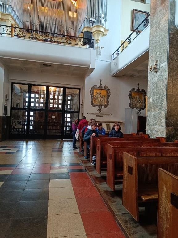 Grupa osób znajduje się w kościele, gdzie część z nich siedzi w ławkach, a inni stoją, oglądając wnętrze świątyni z widocznymi organami na chórze oraz bocznymi obrazami.