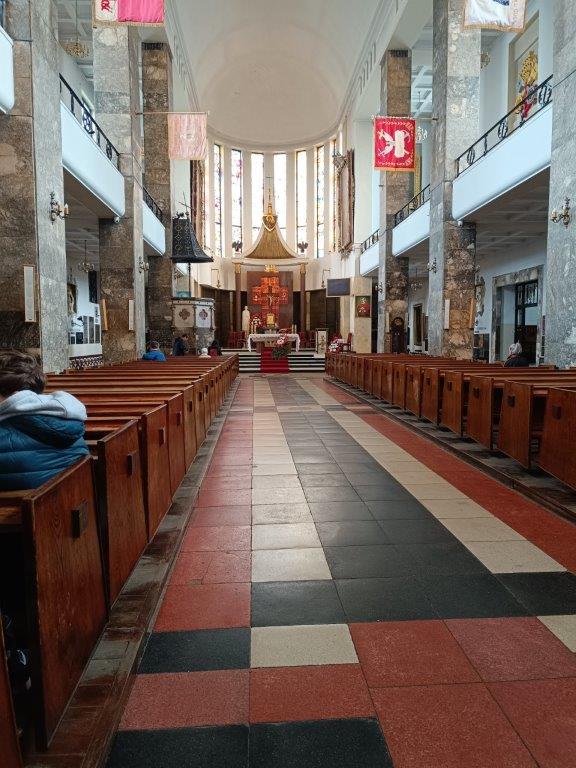Zdjęcie, które przedstawia wnętrze kościoła z długą nawą prowadzącą do ołtarza, po bokach której znajdują się rzędy drewnianych ławek dla wiernych. Charakterystyczny jest tutaj wzorzysty, czarno-biało-czerwony chodnik na posadzce oraz duże filary i wysokie okna, a także proporce wiszące wzdłuż nawy.