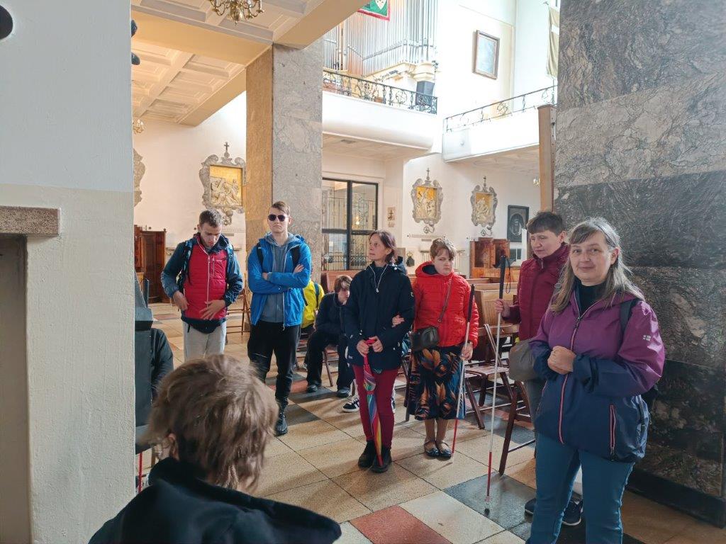 Zdjęcie przedstawia grupę osób znajdującą się w kościele lub sanktuarium. Kilka osób stoi, w tym jedna kobieta, która uśmiecha się do obiektywu. W tle widoczne są elementy wystroju świątyni, takie jak marmurowe filary, obrazy i balkon z organami.