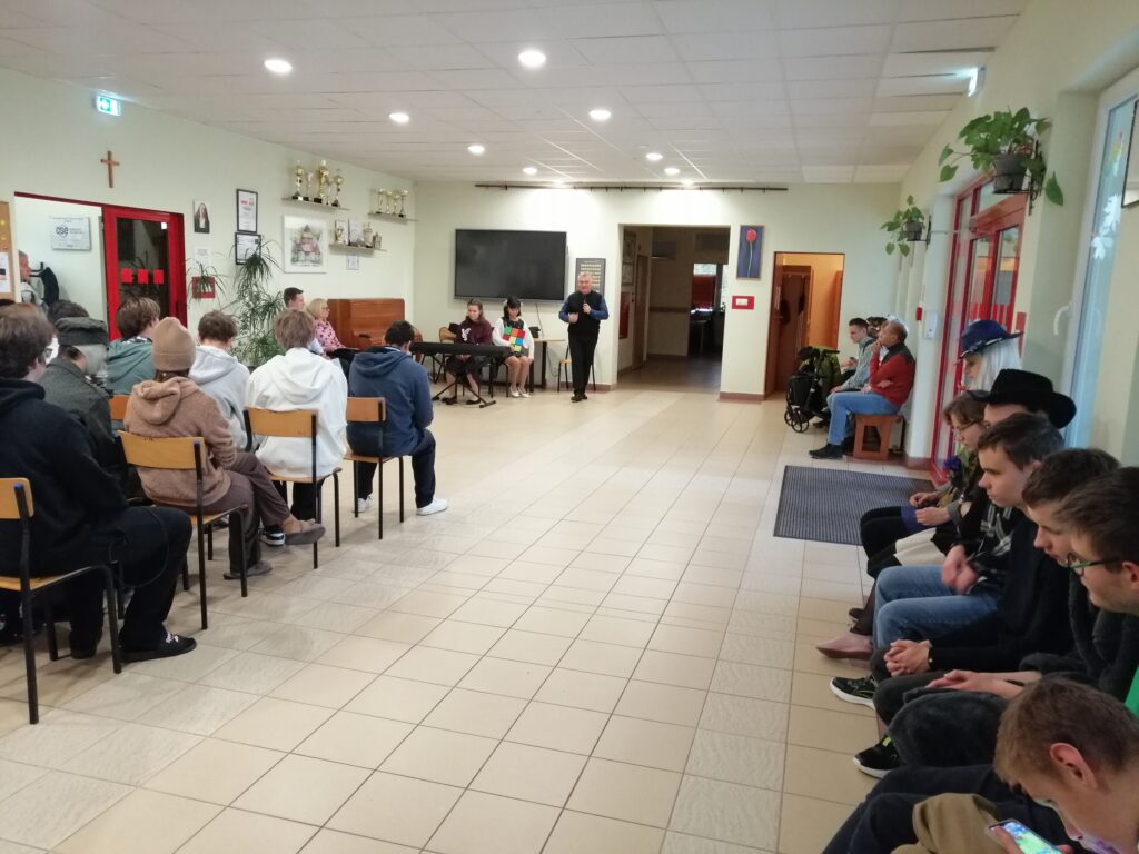 Zdjęcie przedstawia Pana dyrektora szkoły ponadpodstawowej, który wita uczniów zgromadzonych na korytarzu, z okazji Dnia Edukacji Narodowej. Dyrektor stoi w głębi pomieszczenia, przemawiając do młodzieży siedzącej na krzesłach.