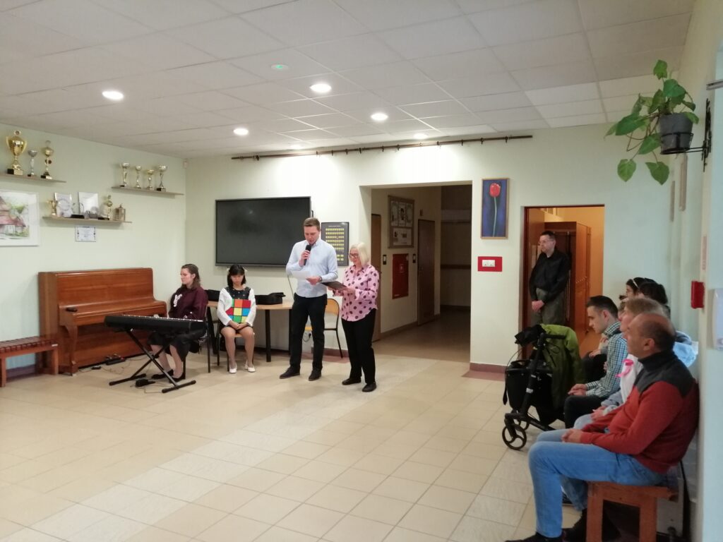 Wychowawcy swoich klas czytają wesołe rady dla nowych uczniów. Uczniowie zgromadzeni na korytarzu słuchają z uwagą. 