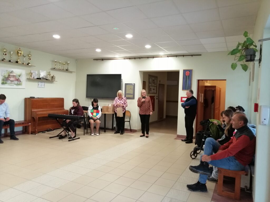 Na zdjęciu widoczne są dwie nauczycielki stojące centralnie, z kartkami w rękach, wygłaszające rozstrzygnięcie konkursu "Popraw poetę" na szkolnym korytarzu. W tle widoczne są instrumenty muzyczne i osoby obserwujące wydarzenie.