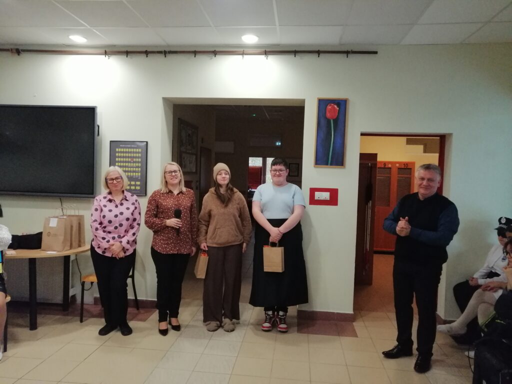 Zdjęcie przedstawia scenę z uroczystości szkolnej na korytarzu, gdzie w centrum stoją uśmiechnięci laureaci konkursu z nagrodami stojącymi obok nauczycieli i dyrektora z uśmiechem na twarzy. 