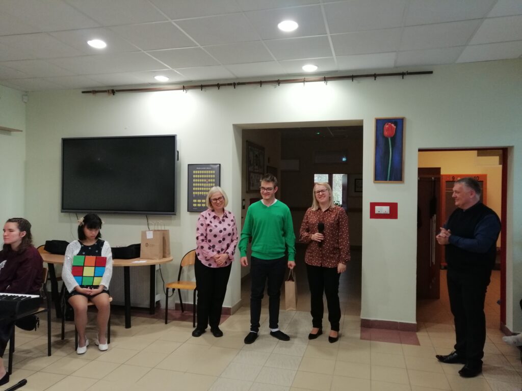 Zdjęcie przedstawia scenę z uroczystości szkolnej na korytarzu, gdzie w centrum stoi  laureat konkursu z nagrodą stojącym obok nauczycieli i dyrektora z uśmiechem na twarzy. 