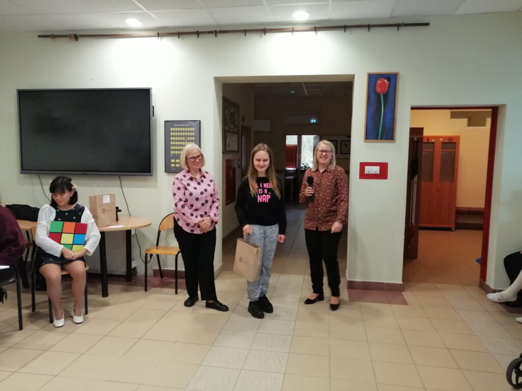 Zdjęcie przedstawia scenę z uroczystości szkolnej na korytarzu, gdzie w centrum stoi  laureatka konkursu z nagrodą stojącą obok nauczycieli z radością na twarzy.