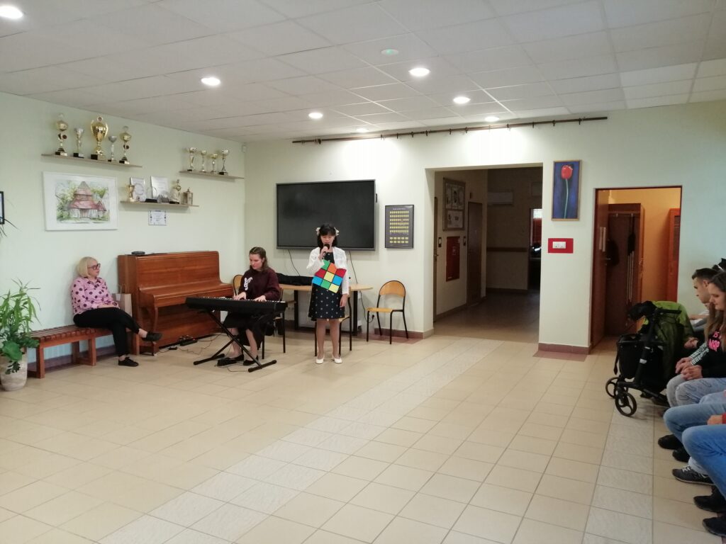 Zdjęcie ukazuje występ artystyczny. Centralnie na planie widać dwie uczennice – jedna gra na keyboardzie, a druga śpiewa do mikrofonu, trzymając w rękach dużą kolorową kostkę. Z lewej strony siedzi nauczycielka, a z prawej widać siedzących uczniów.