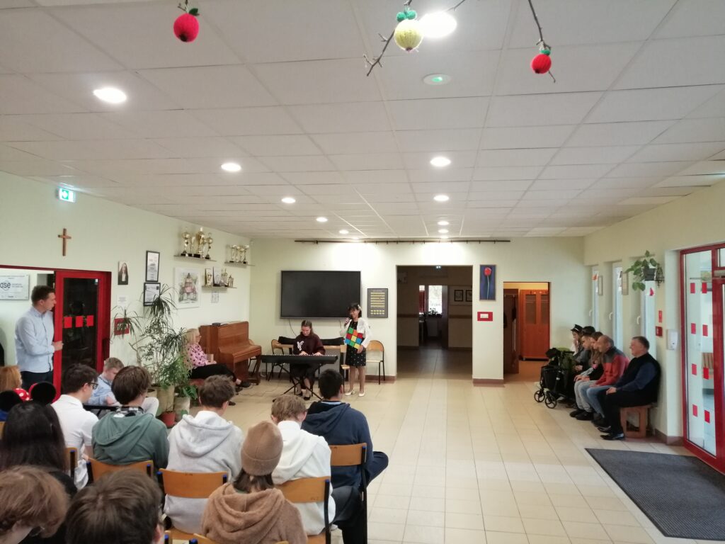 Zdjęcie ukazuje występ artystyczny. Centralnie na planie widać dwie uczennice – jedna gra na keyboardzie, a druga śpiewa do mikrofonu, trzymając w rękach dużą kolorową kostkę. W tle widać uczniów i nauczycieli.