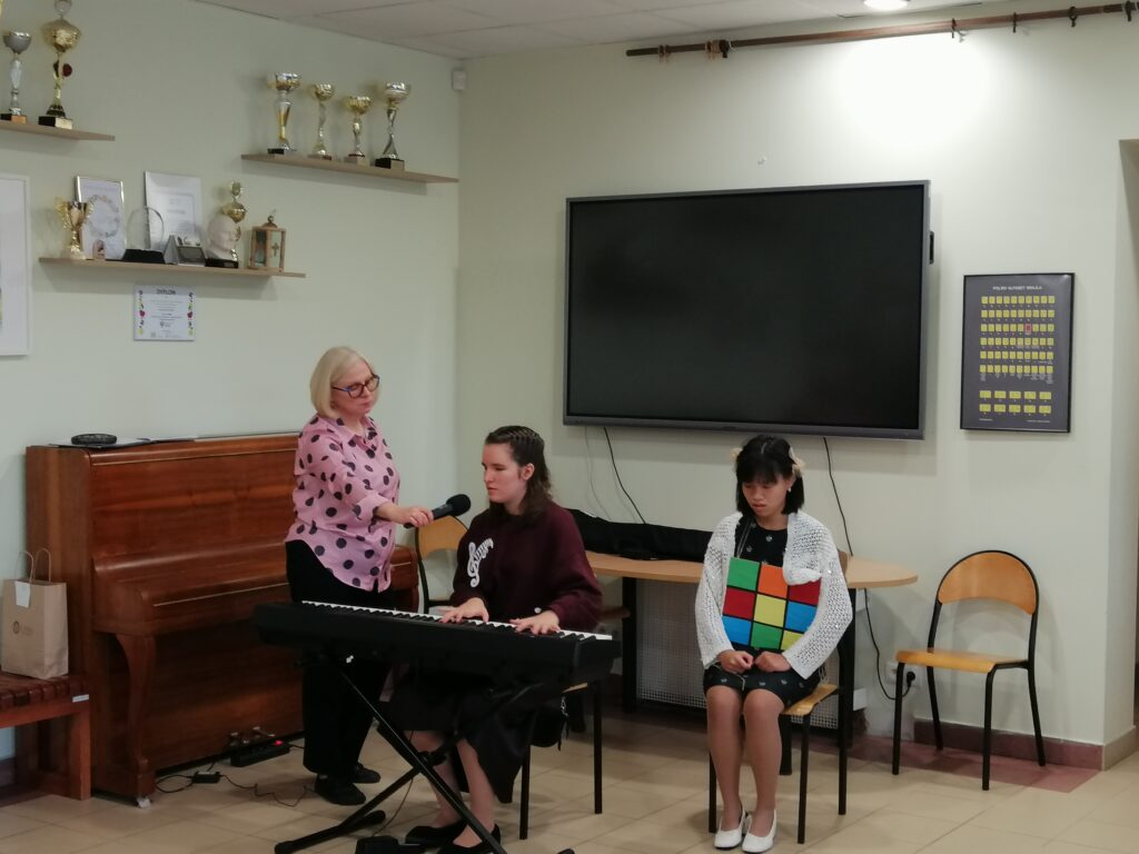 Zdjęcie przedstawia zbliżenie na część artystyczną uroczystości szkolnej, gdzie trzy osoby są zaangażowane w występ. Nauczycielka podaje mikrofon uczennicy grającej na keyboardzie, a obok siedzi inna uczennica, trzymająca dużą, kolorową kostkę. W tle widać pianino, duży ekran oraz półkę z trofeami.