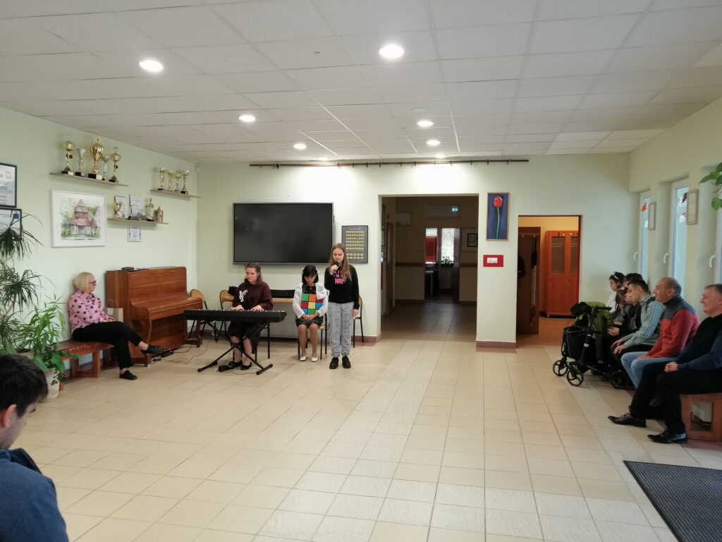 Zdjęcie przedstawia uczennice podczas występu artystycznego na szkolnym korytarzu z okazji, Dnia Edukacji Narodowej. Jedna dziewczyna stoi i śpiewa do mikrofonu, a druga gra na keyboardzie. 