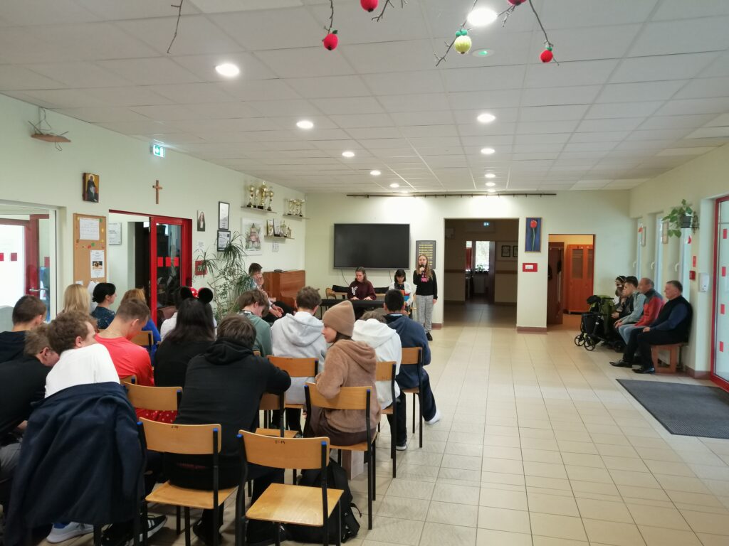 Zdjęcie przedstawia uczennice podczas występu artystycznego na szkolnym korytarzu z okazji, Dnia Edukacji Narodowej. Jedna dziewczyna stoi i śpiewa do mikrofonu, a druga gra na keyboardzie. W tle uczestnicy słuchają piosenki.