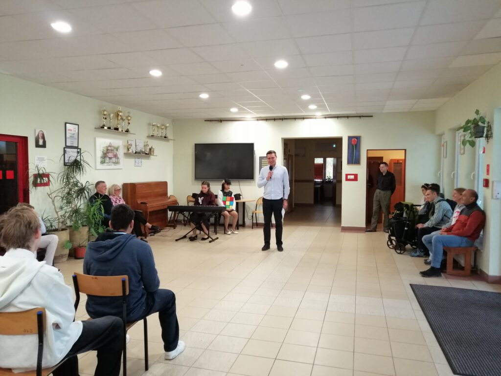 Zdjęcie przedstawia nauczyciela, przemawiającego do zgromadzonych uczniów na korytarzu z widocznymi trofeami na półce oraz instrumentem muzycznym w tle.